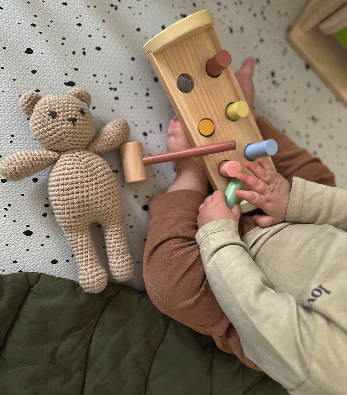 Child playing with a wooden hammer and block set on terrazzo play mat with a teddy bear nearby.